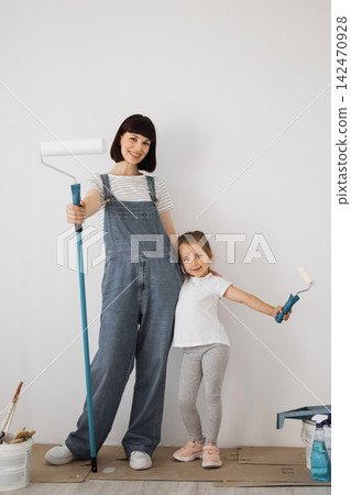 A smiling mother and her young daughter stand side-by-side, holding paint rollers, ready to redecorate a room A smiling mother and her young daughter stand side-by-side, holding paint rollers, ready to redecorate a room 142470928