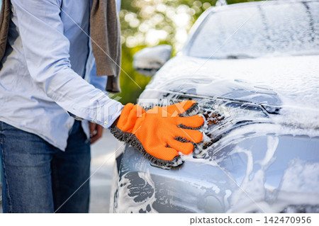 A man in jeans and a blue shirt washes his car with a bright orange microfiber mitt, creating a lot of suds A man in jeans and a blue shirt washes his car with a bright orange microfiber mitt, creating a lot of suds 142470956