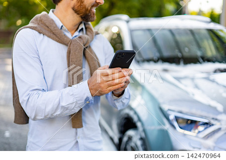 A smiling caucasian man in a light blue shirt and sweater vest uses his smartphone while standing next to a car covered in soap suds A smiling caucasian man in a light blue shirt and sweater vest uses his smartphone while standing next to a car covered in soap suds 142470964