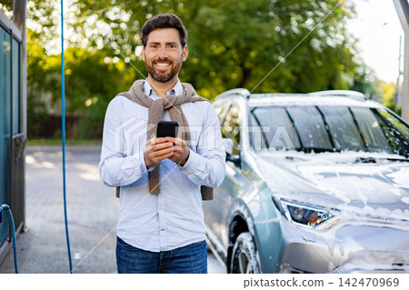 A smiling Caucasian man stands with his smartphone in hand next to a car covered in soap suds at an outdoor car wash station A smiling Caucasian man stands with his smartphone in hand next to a car covered in soap suds at an outdoor car wash station 142470969