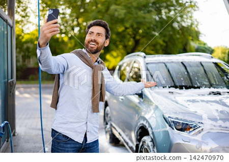 A smiling caucasian man holds up his phone to take a selfie while washing his car with soap and water at an outdoor car wash station A smiling caucasian man holds up his phone to take a selfie while washing his car with soap and water at an outdoor car wash station 142470970