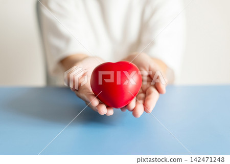 Close up of hands holding red heart on blue background for charity and healthcare concept Close up of hands holding red heart on blue background for charity and healthcare concept 142471248