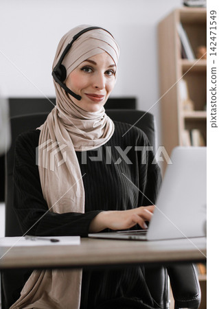 A smiling woman wearing a hijab and headset works on a laptop at her desk, representing customer support A smiling woman wearing a hijab and headset works on a laptop at her desk, representing customer support 142471549