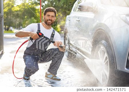 A smiling man in overalls is using a high-pressure washer to clean a car outdoors on a sunny day, creating a lot of foam and spray A smiling man in overalls is using a high-pressure washer to clean a car outdoors on a sunny day, creating a lot of foam and spray 142471571