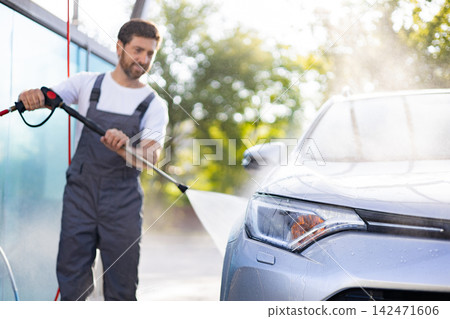 A man in overalls uses a pressure washer to clean a silver car outdoors with water spraying and bokeh background A man in overalls uses a pressure washer to clean a silver car outdoors with water spraying and bokeh background 142471606