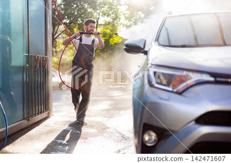 A man in overalls uses a high-pressure washer to clean a car outdoors on a sunny day, creating a lot of mist and spray 142471607
