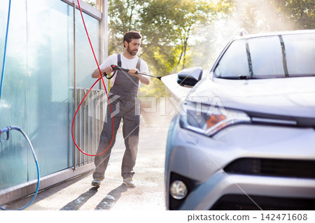 A man in overalls uses a high-pressure washer to clean a silver car outdoors on a sunny day A man in overalls uses a high-pressure washer to clean a silver car outdoors on a sunny day 142471608