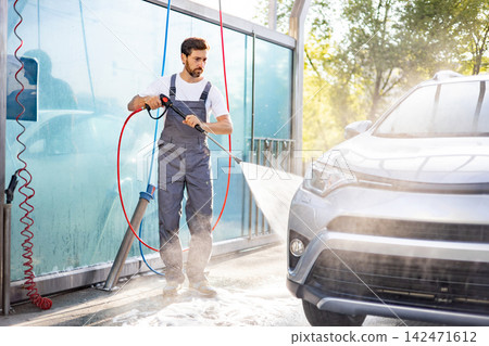 A man in overalls uses a high-pressure water jet to clean a silver car at an outdoor car wash station A man in overalls uses a high-pressure water jet to clean a silver car at an outdoor car wash station 142471612