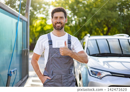 A smiling caucasian man in overalls gives a thumbs up gesture while standing next to a car at a car wash A smiling caucasian man in overalls gives a thumbs up gesture while standing next to a car at a car wash 142471620