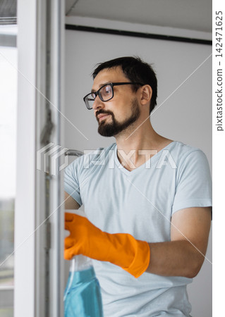 A man wearing orange gloves and glasses sprays a window with a cleaning solution, preparing to wash it A man wearing orange gloves and glasses sprays a window with a cleaning solution, preparing to wash it 142471625
