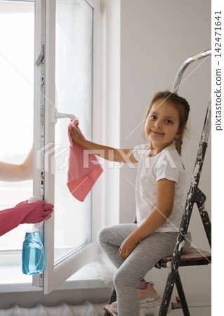 A young girl sits on a ladder and wipes a window with a pink cloth while another person sprays cleaner A young girl sits on a ladder and wipes a window with a pink cloth while another person sprays cleaner 142471641