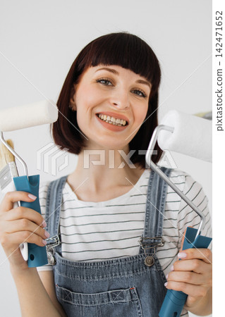 A smiling woman in denim overalls and a striped shirt holds painting tools, ready for a home improvement project A smiling woman in denim overalls and a striped shirt holds painting tools, ready for a home improvement project 142471652