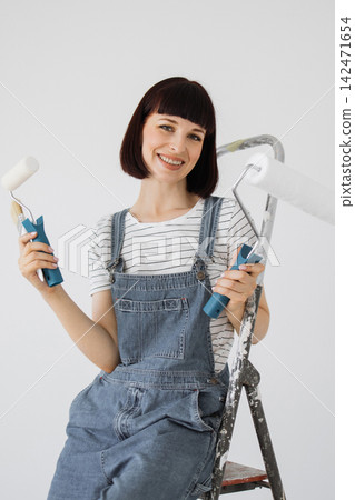 A smiling woman in denim overalls holds paint rollers and a brush while sitting on a ladder, ready for home improvement A smiling woman in denim overalls holds paint rollers and a brush while sitting on a ladder, ready for home improvement 142471654