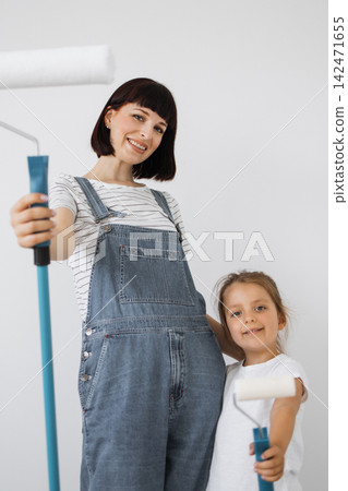 A woman and a young girl in denim overalls smile while holding paint rollers, ready to redecorate a room A woman and a young girl in denim overalls smile while holding paint rollers, ready to redecorate a room 142471655