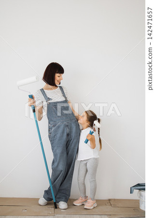 A mother and her young daughter stand side-by-side, holding paint rollers and looking at each other with smiles as they prepare to paint a room A mother and her young daughter stand side-by-side, holding paint rollers and looking at each other with smiles as they prepare to paint a room 142471657