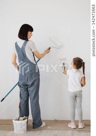 A woman and a young girl stand side-by-side, painting a white wall with rollers, enjoying a home improvement project A woman and a young girl stand side-by-side, painting a white wall with rollers, enjoying a home improvement project 142471668