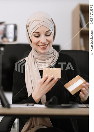 A smiling woman wearing a hijab opens a small cardboard box, possibly for a video review or unboxing A smiling woman wearing a hijab opens a small cardboard box, possibly for a video review or unboxing 142471971