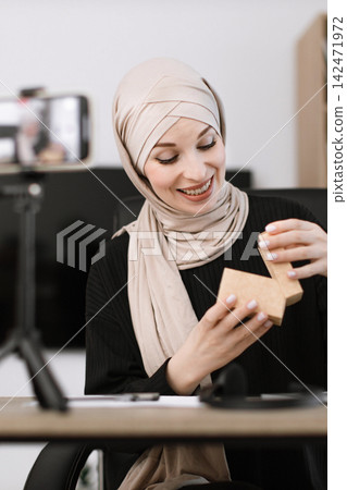 A smiling woman wearing a hijab opens a small cardboard box while recording herself on a smartphone for a live stream or video content A smiling woman wearing a hijab opens a small cardboard box while recording herself on a smartphone for a live stream or video content 142471972