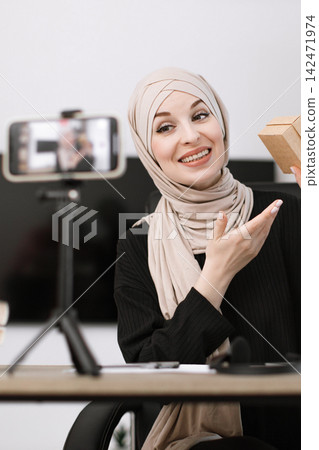 A smiling woman wearing a hijab is recording a video on her smartphone while holding a small gift box, showcasing a product or sharing content online A smiling woman wearing a hijab is recording a video on her smartphone while holding a small gift box, showcasing a product or sharing content online 142471974
