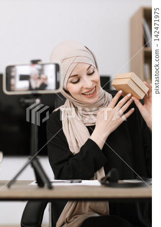 A smiling woman wearing a hijab is recording herself unboxing a small gift box while looking at her phone camera A smiling woman wearing a hijab is recording herself unboxing a small gift box while looking at her phone camera 142471975