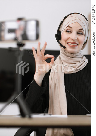 A smiling woman wearing a hijab and headset gives an OK sign while recording a video on her phone A smiling woman wearing a hijab and headset gives an OK sign while recording a video on her phone 142471976