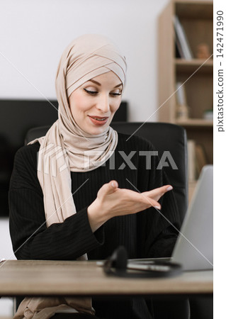 A smiling Muslim woman wearing a hijab is actively engaged in a video call or online meeting on her laptop at her desk A smiling Muslim woman wearing a hijab is actively engaged in a video call or online meeting on her laptop at her desk 142471990