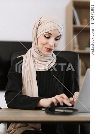 A professional woman wearing a beige hijab and black sweater is focused on her laptop while working at a desk in an office environment A professional woman wearing a beige hijab and black sweater is focused on her laptop while working at a desk in an office environment 142471991