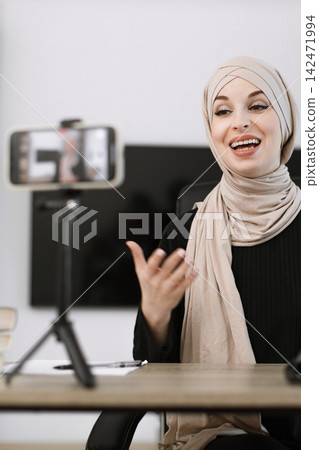 A smiling woman wearing a hijab is recording a video on her smartphone placed on a tripod, gesturing with her hand while sitting at a desk A smiling woman wearing a hijab is recording a video on her smartphone placed on a tripod, gesturing with her hand while sitting at a desk 142471994