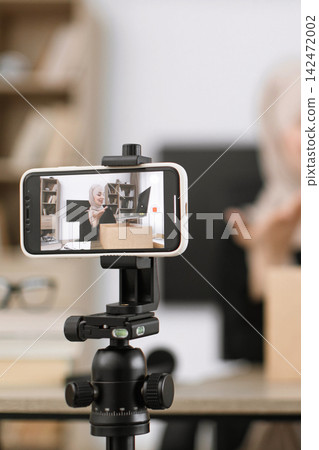 A woman in a hijab records a video on her smartphone, which is mounted on a tripod, showcasing a product box and laptop A woman in a hijab records a video on her smartphone, which is mounted on a tripod, showcasing a product box and laptop 142472002
