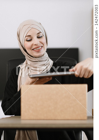 A smiling woman wearing a hijab uses a tablet to scan a cardboard box, likely for inventory or shipping purposes 142472003