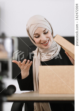 A smiling woman wearing a hijab excitedly opens a cardboard box, revealing its contents A smiling woman wearing a hijab excitedly opens a cardboard box, revealing its contents 142472004