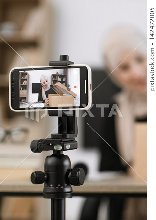 A woman wearing a hijab records herself opening a package on her smartphone mounted on a tripod, creating content for her online channel A woman wearing a hijab records herself opening a package on her smartphone mounted on a tripod, creating content for her online channel 142472005