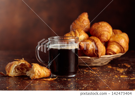 Black coffee and croissants on a brown background. Black coffee and croissants on a brown background. 142473404