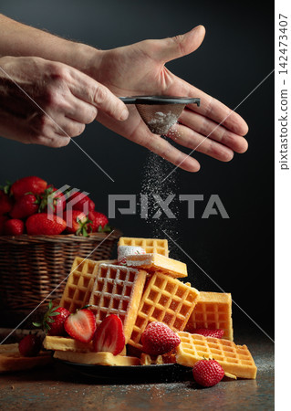 The chef sprinkles powdered sugar on the waffles topped with fresh strawberries. The chef sprinkles powdered sugar on the waffles topped with fresh strawberries. 142473407