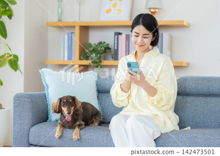 一位女士坐在沙發上看著她的智慧型手機,享受著與狗狗共度的美好時光。 一位女士坐在沙發上看著她的智慧型手機,享受著與狗狗共度的美好時光。 142473501