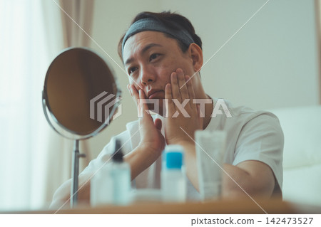 A 40-year-old man, worried about his aging face, touches his cheeks with both hands in front of the mirror and discusses anti-aging care. A 40-year-old man, worried about his aging face, touches his cheeks with both hands in front of the mirror and discusses anti-aging care. 142473527