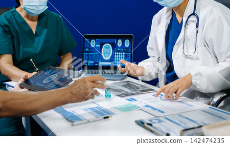 A dedicated medical team discusses patient diagnostics, reviewing brain MRIs and medical charts on digital devices 142474235