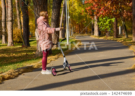 Woman in pink jacket with a scooter points in direction ,autumn park Woman in pink jacket with a scooter points in direction ,autumn park 142474699