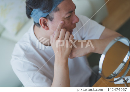 A 40-year-old man worries about aging care, checking the spots and wrinkles on his cheeks with his fingers in front of the mirror. A 40-year-old man worries about aging care, checking the spots and wrinkles on his cheeks with his fingers in front of the mirror. 142474967