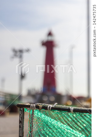 Close-up of green fishing net and rope secured to a weathered metal frame against a blurred waterfront background. 142475735