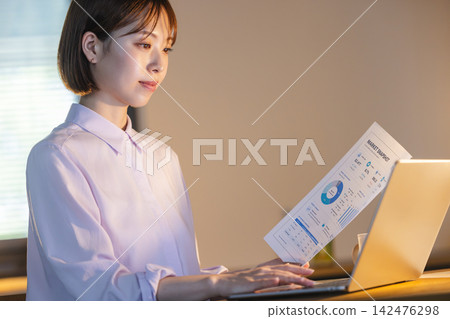 一名女員工正在辦公室的電腦上工作，查看文件中的圖表資料。 142476298
