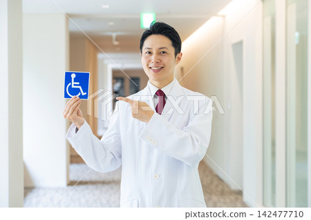 A man in a white coat holding a wheelchair symbol. Doctor, caregiver, welfare, hospital, support, assistance, service, elderly, disabled, medical A man in a white coat holding a wheelchair symbol. Doctor, caregiver, welfare, hospital, support, assistance, service, elderly, disabled, medical 142477710