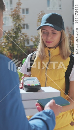 Vertical video. Man open door to a pizza delivery woman wearing yellow thermal backpack with POS wireless terminal for smartphone paying and recieves cardboard boxes of pizza. Food delivery concept. 142478423