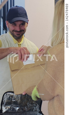 Vertical video. Woman opens door to food delivery man taking order out of thermal insulated bag and giving it to female customer. View from the apartment. Inside shot. Food delivery concept. Vertical video. Woman opens door to food delivery man taking order out of thermal insulated bag and giving it to female customer. View from the apartment. Inside shot. Food delivery concept. 142478469