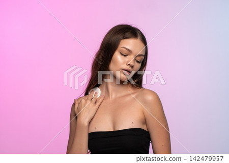 Young woman with closed eyes using a cotton pad for cleansing her neck, prioritizing skincare and self-care 142479957
