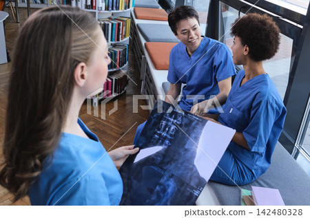 Group of three multiracial intern doctors seated in the campus library assessing spinal column radiography for posture disorders Group of three multiracial intern doctors seated in the campus library assessing spinal column radiography for posture disorders 142480328