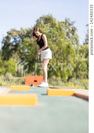 Mini golf activity takes place on a sunny day in a park with a young woman focusing on her shot Mini golf activity takes place on a sunny day in a park with a young woman focusing on her shot 142480335