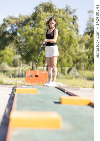 Young woman enjoys mini golf at a recreational park on a sunny day Young woman enjoys mini golf at a recreational park on a sunny day 142480341