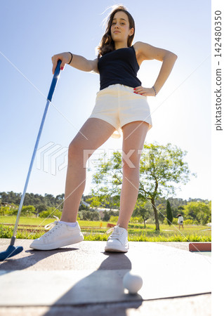Young girl stands confidently with a golf club in an outdoor setting during daytime while preparing for a swing Young girl stands confidently with a golf club in an outdoor setting during daytime while preparing for a swing 142480350