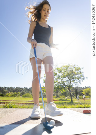 Young girl practices mini golf on a sunny day in a park with green trees and blue sky in the background Young girl practices mini golf on a sunny day in a park with green trees and blue sky in the background 142480367